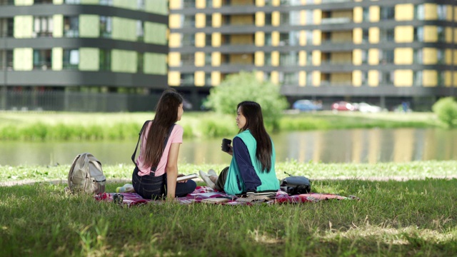 两个不同的女学生在池塘附近的草坪上放松和交谈。喝咖啡的亚洲女孩，拿着书的白人女孩视频素材