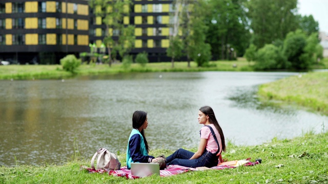 两个大学女生坐在公园池塘边的毯子上聊天。放松的学生，亚洲和高加索人，课后在自然中聊天，周围的书籍，笔记本电脑和背包视频素材