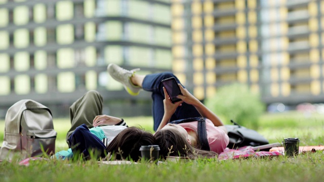 两个女同学下课后在校园的草坪上休息。女孩们躺在毯子上，浏览智能手机，聊天视频素材