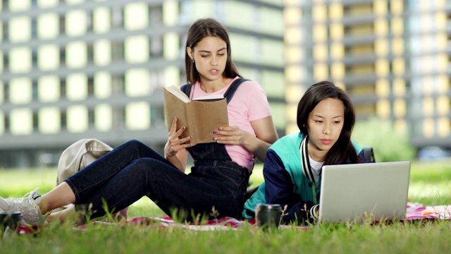 两个不同的女学生在草地上的毯子上一起做大学项目。一个白人女孩在看书，她的亚洲朋友在电脑上打字。学生讨论的话题视频素材