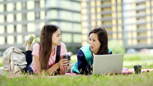 两个不同的女学生躺在草地上的毯子上，讨论着家庭作业。放松的大学朋友喝咖啡和微笑，打开笔记本电脑在亚洲女孩面前视频素材