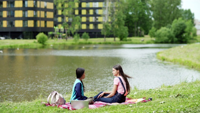 两个不同的女学生坐在公园池塘边的毯子上聊天。放松的大学朋友，亚洲和高加索人，喝咖啡和聊天在自然课后视频素材