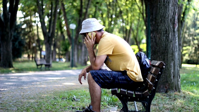 戴着帽子和太阳镜的人坐在公园的长凳上打电话。夏天的一天，在公园的树下，一名男子一边打电话一边放松视频素材