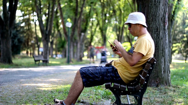 戴着帽子和太阳镜的男人正在公园的长凳上使用智能手机。夏日的某天，一名男子在公园的树下放松时使用智能手机视频素材