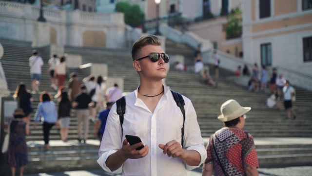 年轻英俊的游客在市中心迷路了。穿着衬衫，背着旅游背包的男子正在用智能手机查看在线手机应用。戴墨镜的人环顾四周，用导航程序确定正确的位置视频素材