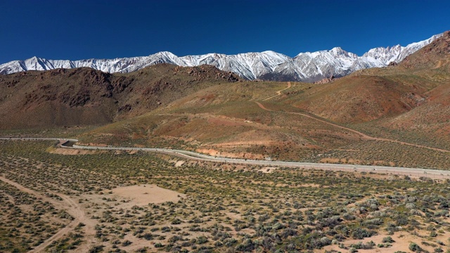 空中下降:一条蜿蜒的道路在干旱的山丘和平原与高积雪覆盖的山脉-阿拉巴马山，加利福尼亚视频素材