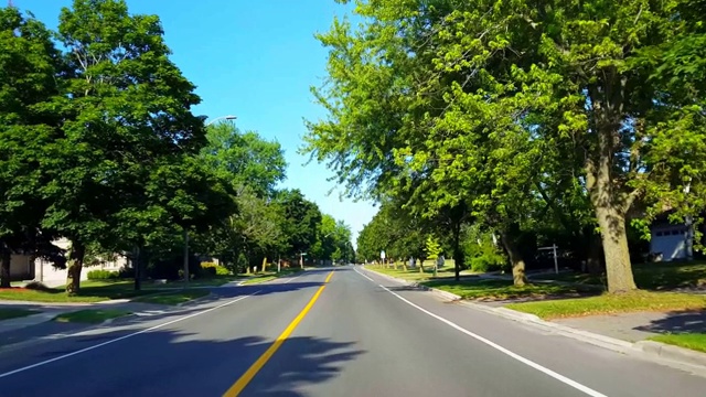 夏季驾驶绿树成荫的住宅城市道路。司机视角沿着美丽的阳光郊区街道。视频素材