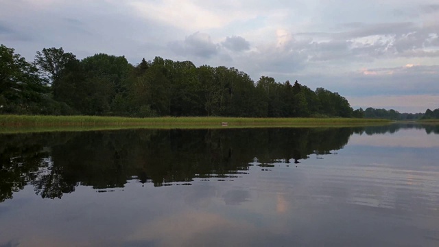 在宁静的夏夜，美丽的自然景观。湖岸与绿色的树木和植物反射在水晶干净镜面水面。天空乌云密布。视频素材