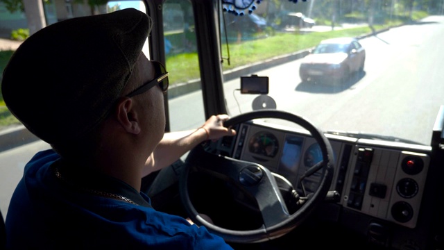 在阳光明媚的日子里，在城市道路上行驶的货车司机在车厢里享受旅程。戴着帽子和太阳镜的男人控制着他的卡车，专注地看着道路。内景慢镜头视频素材