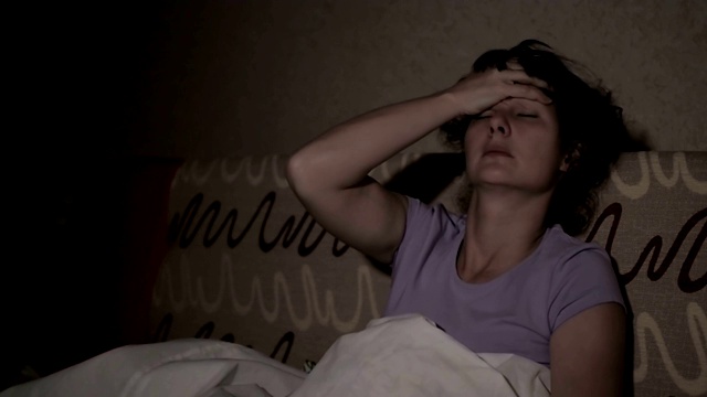 一位年轻女子正遭受着严重的头痛。感冒发烧。一个生病的女孩晚上躺在沙发上，盖着毯子，用手摸着自己的头。视频素材