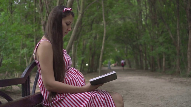 一个怀孕的女孩坐在公园的长椅上。一个穿着白红条纹连衣裙的女孩正在兴致勃勃地看书。视频素材