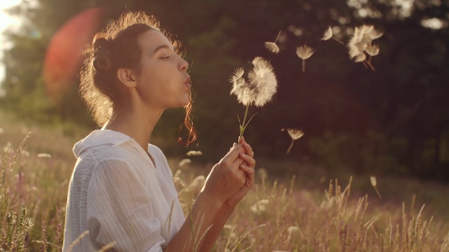 美丽的年轻女子在夏日日落的麦田里吹蒲公英。美夏理念视频素材