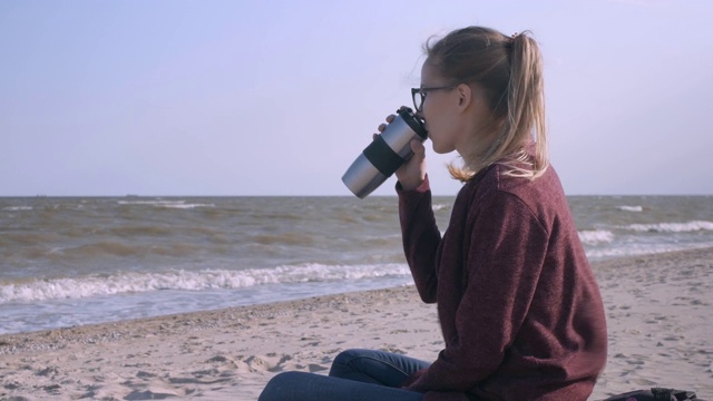 英俊的白人女人坐在海边的海滩，享受美好的一天，喝咖啡(或茶)。风吹在头发上。视频素材