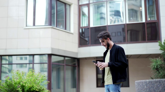 一名年轻男子拿着外卖咖啡杯走在大街上，却收到智能手机上的短信。男人停下来看手机短信，面带微笑视频素材