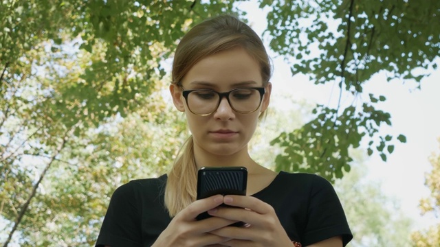 戴眼镜的漂亮白人女人用黑色手机坐在城市公园的长椅上视频素材