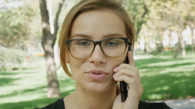 快乐的年轻白种女人戴眼镜在谈论黑色手机，而坐在长椅上的城市公园在夏天的一天。特写肖像。视频素材