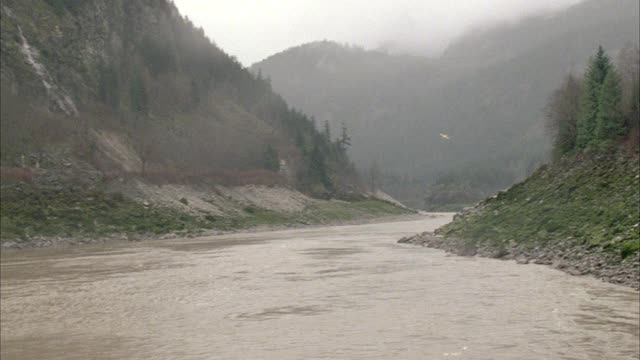 宽角度的河谷与山脉在fg和bg。松树和悬崖可见。黄色单引擎浮筒水上飞机飞越河流或小溪。多云。系列。视频素材