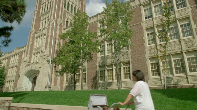 广角三层砖砌建筑，可以是高中或学院，大学，常春藤盟校校园。女人推着婴儿车走过去。视频素材