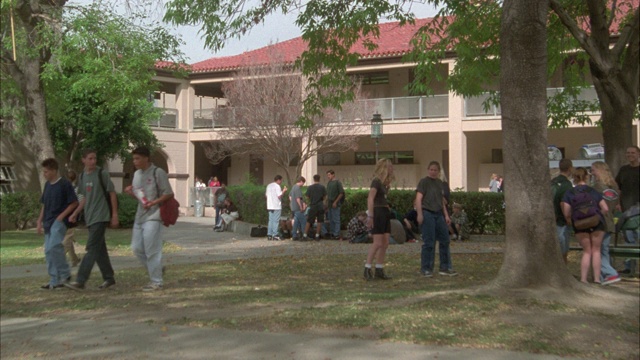 广角的学生，青少年背着书包走过中学或高中的庭院区域。两层建筑，西班牙风格的红瓦屋顶在bg。学生们围坐在院子中间的长椅上。视频素材