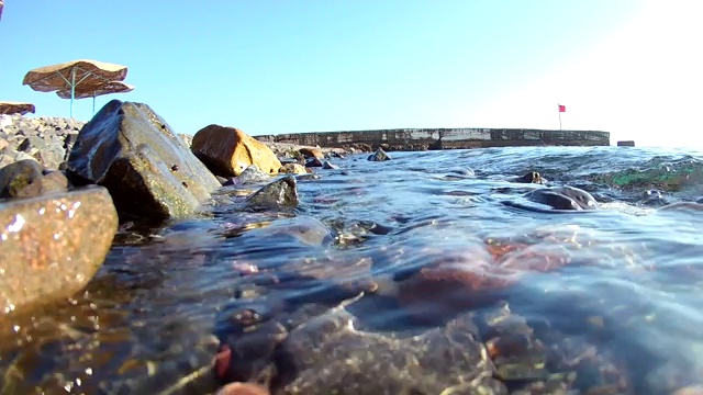 海浪翻滚到海岸岩石海岸的海滩上，有茅草伞，你可以看到一个码头红旗，水下的看法。视频素材