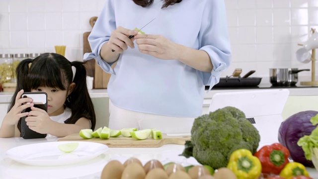 幸福的亚洲家庭妈妈正在准备蔬菜和水果，女儿在家里的厨房玩智能手机。健康饮食和晨间活动理念视频素材