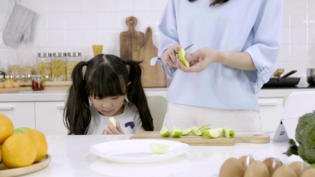 幸福的亚洲家庭妈妈正在准备蔬菜和水果，女儿在家里的厨房玩智能手机。健康饮食和晨间活动理念视频素材