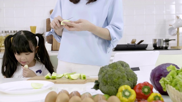 幸福的亚洲家庭妈妈正在准备蔬菜和水果，女儿在家里的厨房玩智能手机。健康饮食和晨间活动理念视频素材