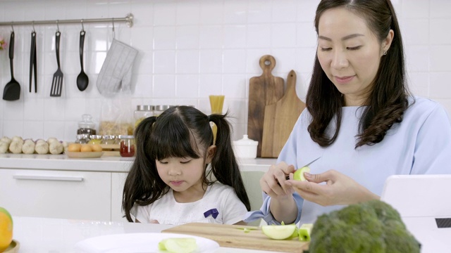 幸福的亚洲家庭妈妈正在准备蔬菜和水果，女儿在家里的厨房玩智能手机。健康饮食和晨间活动理念视频素材