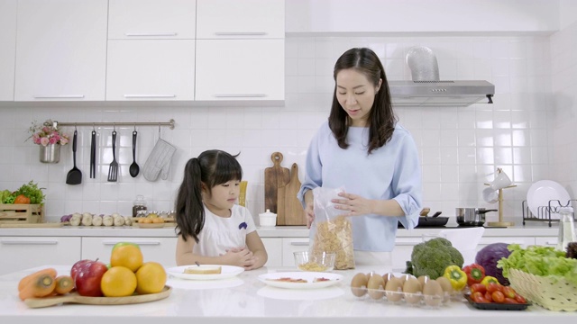 快乐的亚洲家庭妈妈看小女孩正在吃早餐，麦片和牛奶在家里的厨房。健康食品理念为了增强身体的力量视频素材