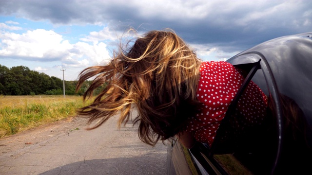 深色头发的女孩探出车窗，在乡村公路上骑车，享受旅行。年轻女子伸出她的头在移动的汽车和她的长棕色头发在风中吹拂。特写慢动作视频素材