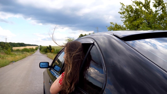 美丽的女人探出车窗，享受旅行，而骑在乡村道路。年轻的女孩看着窗外移动的汽车和她的棕色长发在风中吹拂。慢动作视频素材