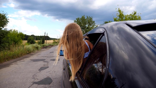 在阴天的乡村道路上，一个小孩看着窗外的汽车。小女孩探出车窗，长长的金发在风中飘动。后视视频素材