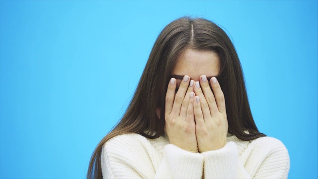 年轻漂亮的女孩站在蓝色的背景上。在这段时间里，她穿着一件白色的暖和的毛衣。脸，她的手，然后打开它和微笑。视频素材