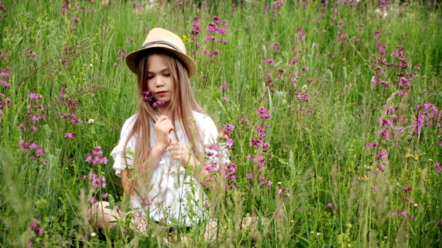 小女孩戴着帽子坐在草地上捧着鲜花。孩子笑了，靠近一点视频素材