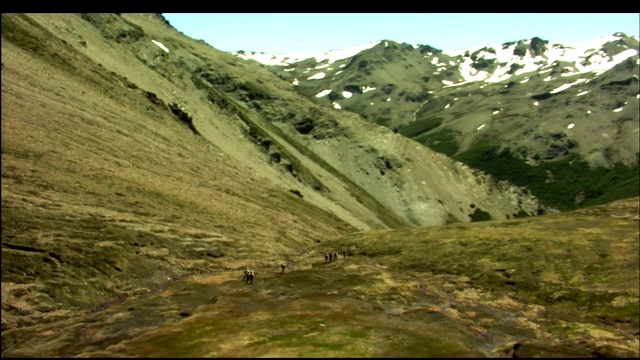 徒步旅行者在白雪覆盖的山峰下沿着干涸的河床行走。视频素材