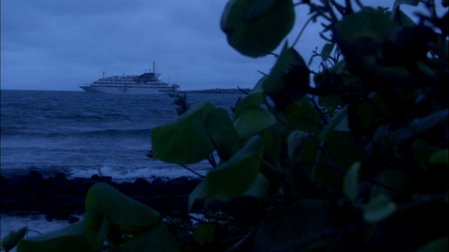 海浪缓缓地滚向郁郁葱葱的海岸线，一艘抛锚停泊的船隐约出现在背景中。视频素材