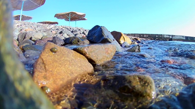 海浪翻滚到海岸岩石海岸的海滩上，有茅草伞，你可以看到一个码头红旗，水下的看法。慢动作视频素材