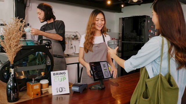 年轻的亚洲女咖啡师向亚洲顾客解释零浪费折扣促销，在咖啡馆的柜台吧台买咖啡时自己带咖啡杯，慢镜头视频素材