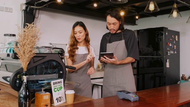 年轻的亚洲咖啡师男女拿着数字平板电脑在咖啡馆的柜台边交谈和计划他们的工作视频素材