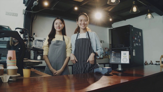 两名年轻美丽的亚洲女咖啡师穿着围裙，微笑着站在咖啡馆的吧台边交谈。咖啡店和小型咖啡店的概念。缓慢的运动。视频素材