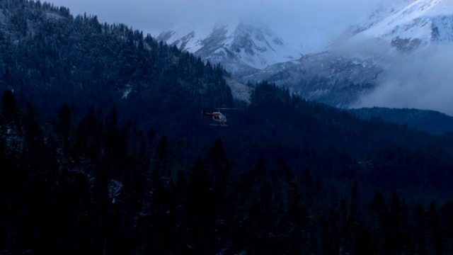 一架直升飞机飞过白雪皑皑、森林覆盖的群山。视频素材