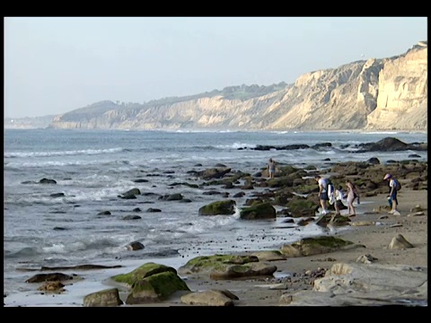 一小群人在海滩上散步时，海浪冲刷着布满岩石的海岸线。视频素材