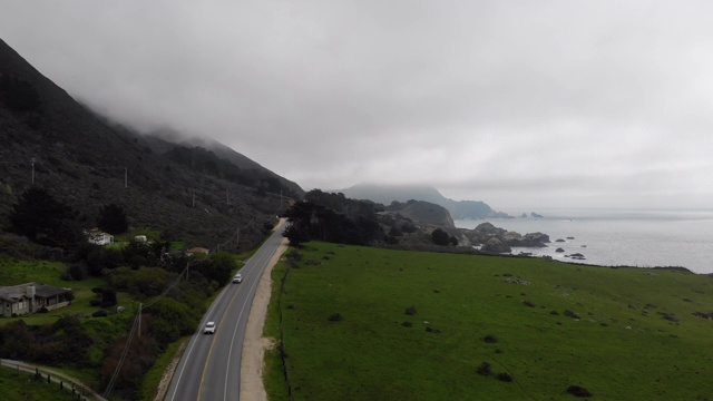 慢慢飞过一条公路，旁边是大山和草地，有悬崖俯瞰大海。汽车慢慢驶过。乌云笼罩着山顶视频下载