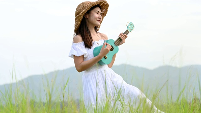 亚洲快乐的女人在白色的衣服弹奏尤克里里吉他在绿色的草地与山的背景在夏天。音乐家美女放松与乐器。乡村的生活方式视频素材