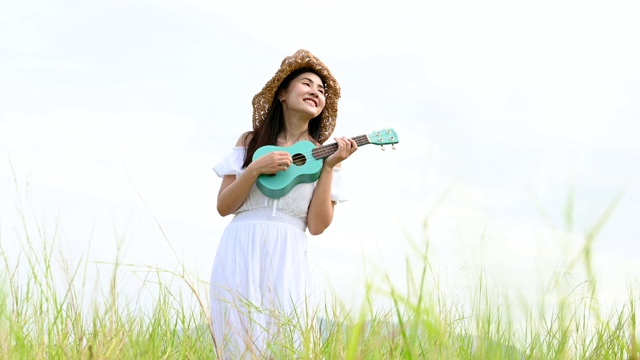 亚洲快乐的女人在白色的衣服弹奏尤克里里吉他在绿色的草地与山的背景在夏天。音乐家美女放松与乐器。乡村的生活方式视频素材