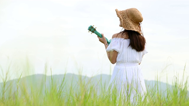 亚洲快乐的女人在白色的衣服弹奏尤克里里吉他在绿色的草地与山的背景在夏天。音乐家美女放松与乐器。乡村的生活方式视频素材