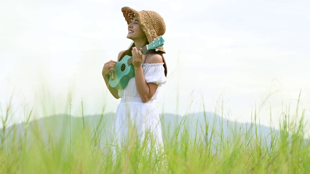 亚洲快乐的女人在白色的衣服弹奏尤克里里吉他在绿色的草地与山的背景在夏天。音乐家美女放松与乐器。乡村的生活方式视频素材