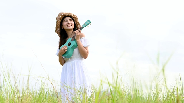 亚洲快乐的女人在白色的衣服弹奏尤克里里吉他在绿色的草地与山的背景在夏天。音乐家美女放松与乐器。乡村的生活方式视频素材