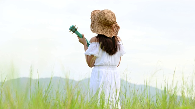 亚洲快乐的女人在白色的衣服弹奏尤克里里吉他在绿色的草地与山的背景在夏天。音乐家美女放松与乐器。乡村的生活方式视频素材