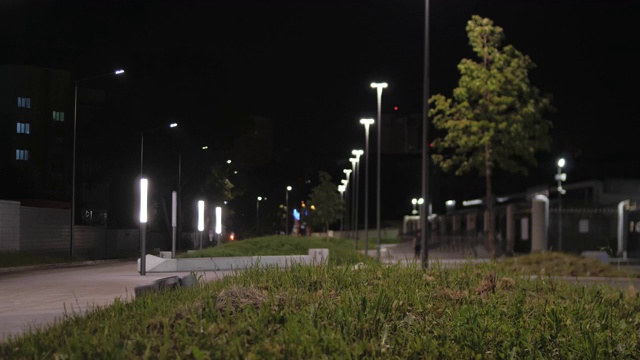 在夏季或春季的夜晚，街道的人行道，绿色的植物和树木和模糊的剪影的人走在人行道上的背景。资料片。城市夜景观视频素材